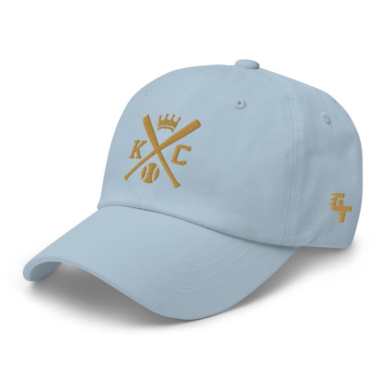 May include: A light blue baseball cap with a gold embroidered logo featuring crossed baseball bats, a baseball, and the letters "KC" with a crown above the letters.
