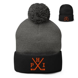 Op de afbeelding: Een antracietgrijze gebreide muts met een zwarte omgeslagen rand en een bijpassende antracietgrijze pompon bovenop. De zwarte rand is voorzien van een oranje geborduurd embleem van twee gekruiste hockeysticks met de letters P, H en I eromheen.