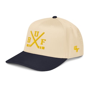 May include: A cream-colored baseball cap with a navy blue brim. The cap features the yellow embroidered letters "BUF" crossed with hockey sticks. A yellow logo is on the side.