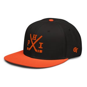 Op de afbeelding: Een zwarte snapback-pet met een oranje platte klep en een oranje knoop bovenop. De voorkant is voorzien van een oranje geborduurde afbeelding van twee gekruiste hockeysticks met de letters P, H en I. Een klein oranje GT-logo staat op het zijpaneel.