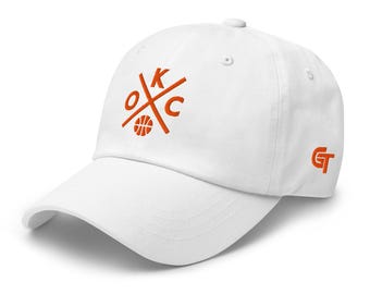 May include: A white baseball cap featuring an embroidered orange logo on the front with the letters OKC arranged around a basketball graphic. The side of the cap displays an orange GT logo. The hat has a curved brim and a structured crown.
