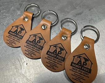 Op de afbeelding: Vier bruin lederen sleutelhangers met zilveren sleutelringen. Elke sleutelhanger heeft een druppelvorm en toont de tekst "HANDY PROS" met een logo en contactgegevens in zwart. De sleutelhangers zijn gemaakt van lichtbruin leer.