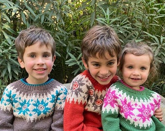May include: Three children wearing colourful knitted sweaters with snowflake patterns. The sweaters are in shades of brown, red, green, and pink.