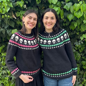 Puede incluir: Dos personas con suéteres negros con diseños de calaveras. Los suéteres tienen cuello redondo y mangas largas. El diseño de calaveras es blanco, con detalles rosas y blancos en un suéter y detalles verdes y blancos en el otro. El fondo es verde.