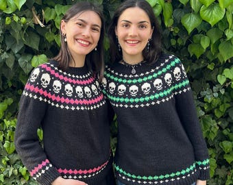 Puede incluir: Dos personas con suéteres negros con diseños de calaveras. Los suéteres tienen cuello redondo y mangas largas. El diseño de calaveras es blanco, con detalles rosas y blancos en un suéter y detalles verdes y blancos en el otro. El fondo es verde.