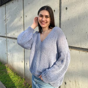 Puede incluir: Un suéter de punto azul claro con cuello en V y mangas largas. El suéter tiene un ajuste holgado y cómodo. La modelo lleva el suéter con vaqueros azules. El fondo es una pared de hormigón.
