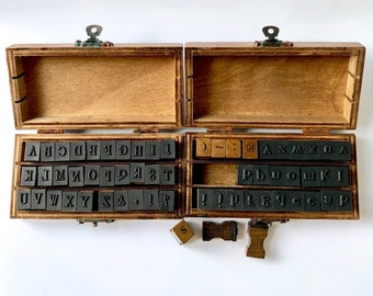 May include: A wooden box containing a set of letter stamps. The stamps are arranged in rows and include letters, numbers, and punctuation marks. The box is open and the stamps are visible. There are also a few extra stamps outside the box.