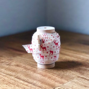 K&ouml;nnte beinhalten: Eine Spule mit wei&szlig;em Stoffband mit einem roten Rentier- und Sternenmuster. Das Band ist um eine Holzspule gewickelt und mit einer Holzklammer befestigt.