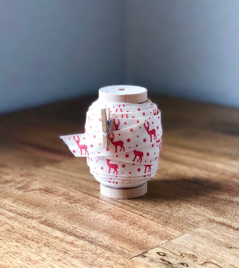 K&ouml;nnte beinhalten: Eine Spule mit wei&szlig;em Stoffband mit einem roten Rentier- und Sternenmuster. Das Band ist um eine Holzspule gewickelt und mit einer Holzklammer befestigt.