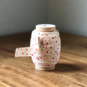 K&ouml;nnte beinhalten: Eine Spule mit wei&szlig;em Stoffband mit rotem Sternmuster. Das Band ist um eine Holzspule gewickelt und mit einer Holzklammer zusammengehalten.