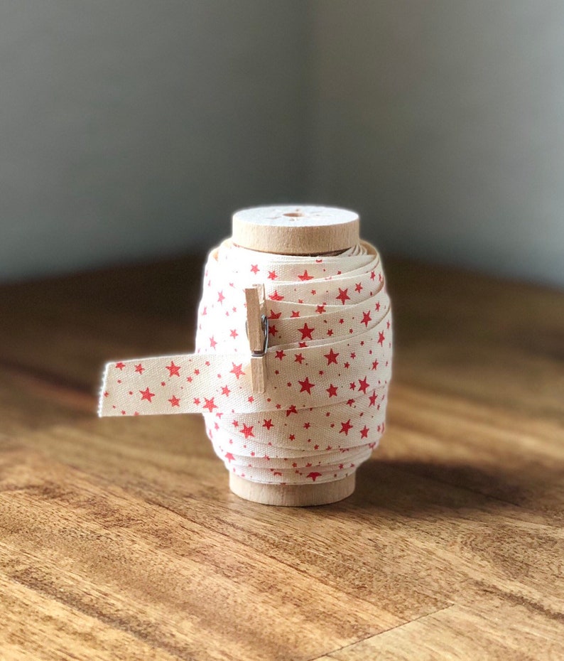 K&ouml;nnte beinhalten: Eine Spule mit wei&szlig;em Stoffband mit rotem Sternmuster. Das Band ist um eine Holzspule gewickelt und mit einer Holzklammer zusammengehalten.