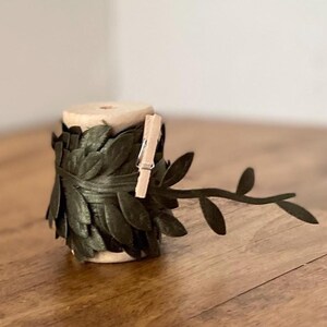 Può includere: Una bobina di ghirlanda di foglie verde scuro con una piccola molletta di legno. La ghirlanda è avvolta attorno a una bobina di legno. Un singolo rametto di foglie si estende dalla bobina.