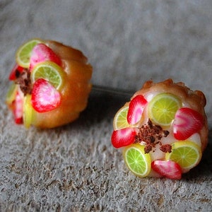 Puede incluir: Un par de mini cupcakes con cobertura de fruta y base de glaseado blanco. Los cupcakes están decorados con fresas rojas, rodajas de lima verde y chispas de chocolate marrón.