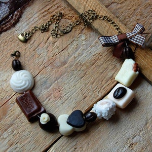 May include: A brown chain necklace with a heart charm and a variety of brown, white and black polymer clay charms shaped like coffee beans, chocolate bars, and swirls. The charms are strung on the chain with a brown and white checked ribbon bow.