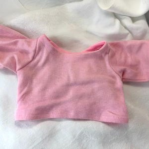 May include: A short-sleeved, light pink t-shirt featuring a wide, rounded neckline and a straight hemline. The garment is constructed from a soft, solid-colored fabric with visible stitching along the sleeve cuffs and the bottom edge of the shirt.