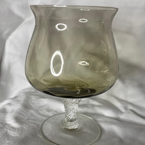 May include: A smoky gray glass goblet with a wide bowl and a clear, textured stem. The goblet has a round base and a decorative, twisted stem. The glass has a slight tint of green at the base. The goblet is designed for beverages.