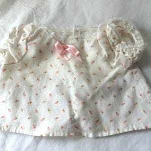 May include: A white short-sleeved top featuring a repeating pattern of tiny pink rosebuds. The garment has gathered puff sleeves with delicate white lace trim at the edges and a small, light pink ribbon bow centered at the neckline.