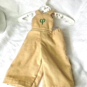 May include: Tan corduroy jumpsuit with a green embroidered logo on the bib, hanging on a white plastic hanger. The jumpsuit has wide legs and adjustable straps. The fabric has vertical ridges.
