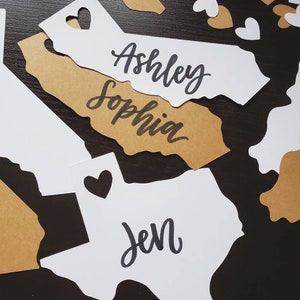 May include: White paper cutouts of the states of California and Texas with the names "Ashley Sophia" and "Jen" written on them. The cutouts are on a dark brown surface with gold paper hearts scattered around them.