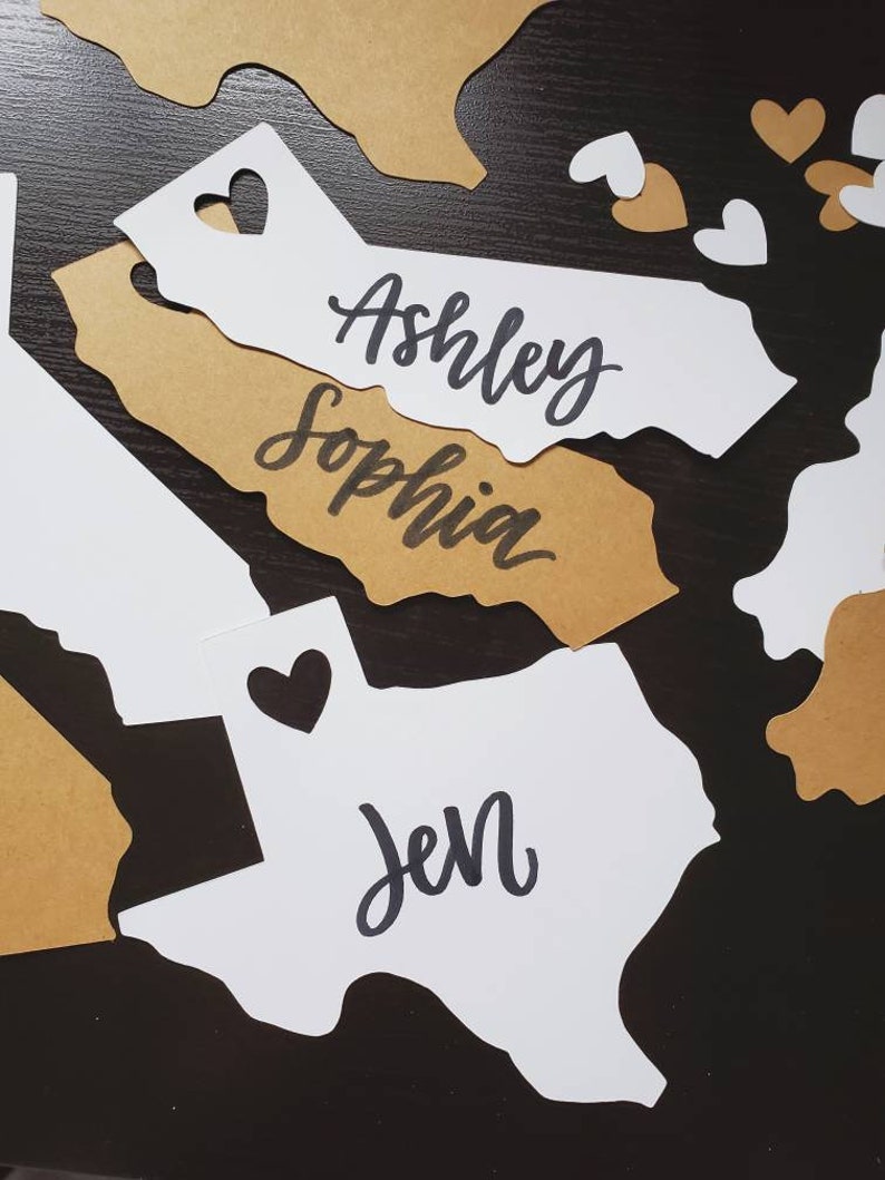 May include: White paper cutouts of the states of California and Texas with the names "Ashley Sophia" and "Jen" written on them. The cutouts are on a dark brown surface with gold paper hearts scattered around them.