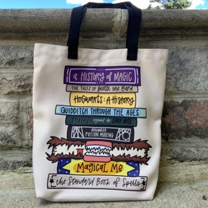 May include: Cream-colored tote bag with black handles. The bag features a stack of illustrated books with titles like "A History of Magic" and "Hogwarts: A History." Below the books is the text "Magical Me" and "the Standard Book of Spells."