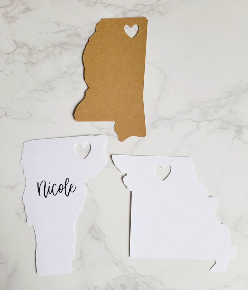 May include: Three white paper state outlines with heart cutouts. The state on the left says "Nicole" and is the state of Vermont. The state on the right is Missouri. The state in the middle is a brown paper outline of Mississippi.