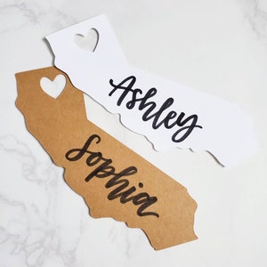 May include: Two place cards shaped like the state of California. One is brown cardstock with the name "Sophia" written in black cursive. The other is white cardstock with the name "Ashley" written in black cursive.