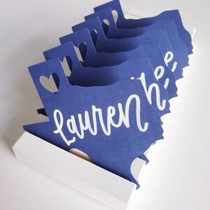 May include: A set of blue paper place cards shaped like the state of Texas with a heart cut out at the top. The name "Lauren" is written in white on the front of the place cards.