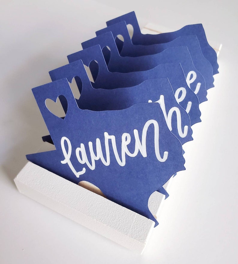 May include: A set of blue paper place cards shaped like the state of Texas with a heart cut out at the top. The name "Lauren" is written in white on the front of the place cards.
