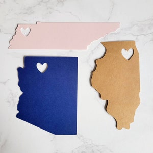 May include: Three state-shaped paper cutouts with heart-shaped holes. The cutouts are in the shapes of Tennessee, Illinois, and Arizona. The Tennessee cutout is pink, the Illinois cutout is brown, and the Arizona cutout is blue.