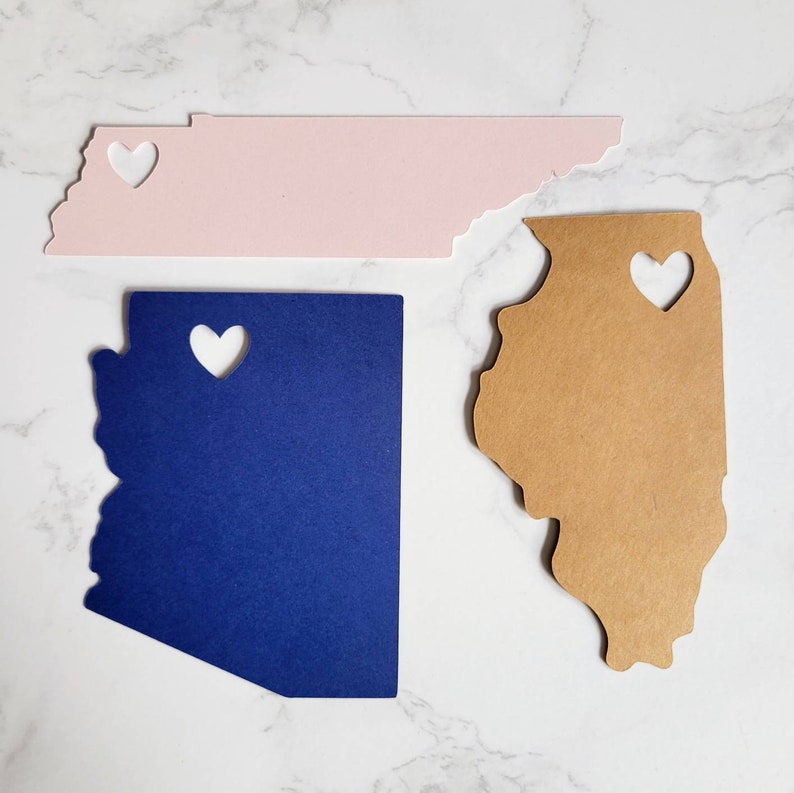 May include: Three state-shaped paper cutouts with heart-shaped holes. The cutouts are in the shapes of Tennessee, Illinois, and Arizona. The Tennessee cutout is pink, the Illinois cutout is brown, and the Arizona cutout is blue.