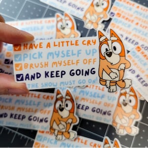 May include: A sticker featuring a cartoon dog with the text "Have a little cry, pick myself up, brush myself off, and keep going. The show must go on!"