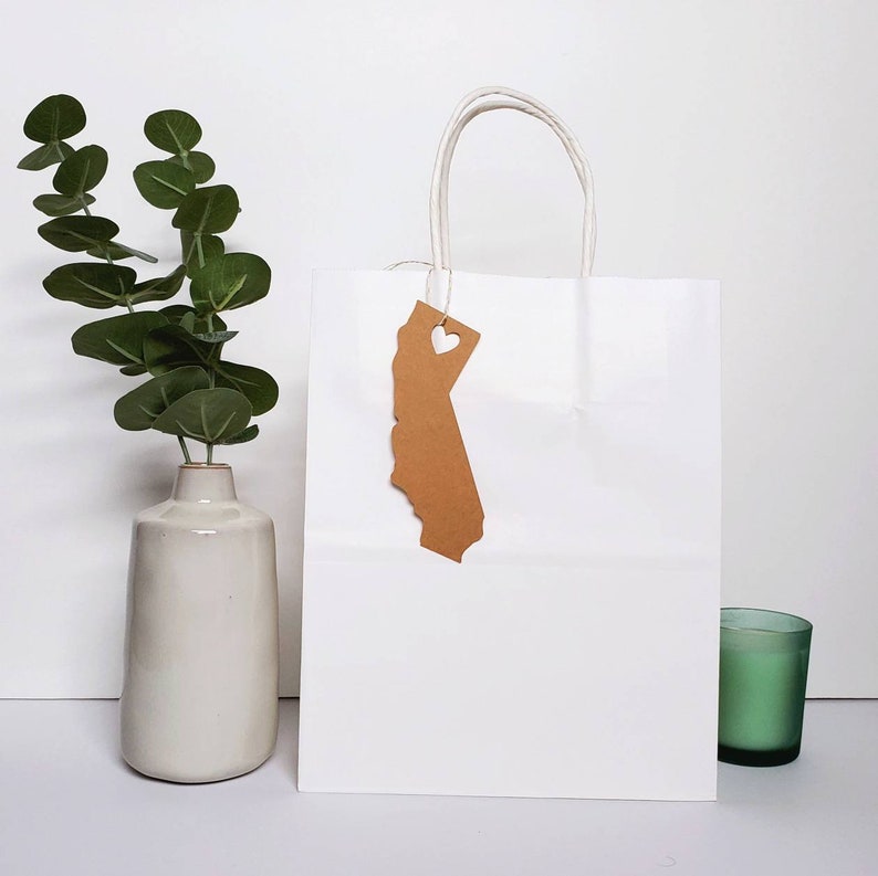 May include: White paper gift bag with a brown paper tag shaped like the state of California with a heart cut out. The bag has white handles and is sitting on a white surface.