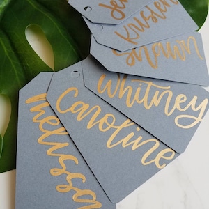 May include: Six gray gift tags with gold lettering. The names Jennifer, Kristen, Shawn, Whitney, Caroline, and Melissa are written on the tags.