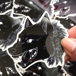 May include: Black and white wolf stickers. The stickers feature a detailed illustration of a snarling wolf's head with sharp teeth and a fluffy mane. The stickers are held by a person's fingers.