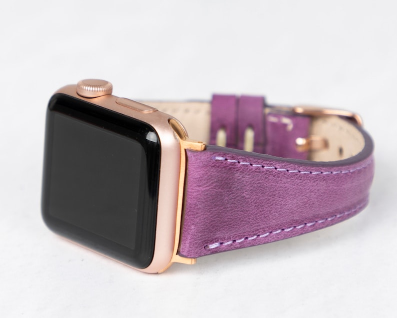 May include: A rose gold Apple Watch with a purple leather band. The watch face is black and the band has white stitching.