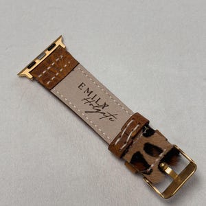 Puede incluir: Una correa de reloj Apple de cuero marr&oacute;n con una hebilla dorada y un dise&ntilde;o de estampado de leopardo. La correa est&aacute; cosida con hilo blanco y tiene el texto "Emily & Wyatt" grabado en la parte posterior.