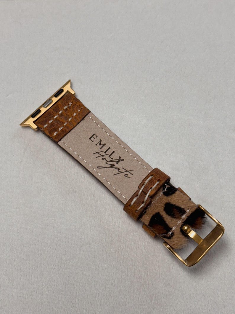 Puede incluir: Una correa de reloj Apple de cuero marr&oacute;n con una hebilla dorada y un dise&ntilde;o de estampado de leopardo. La correa est&aacute; cosida con hilo blanco y tiene el texto "Emily & Wyatt" grabado en la parte posterior.