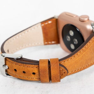May include: A brown leather watch strap with a rose gold Apple Watch attached. The strap has white stitching detail and a silver buckle.