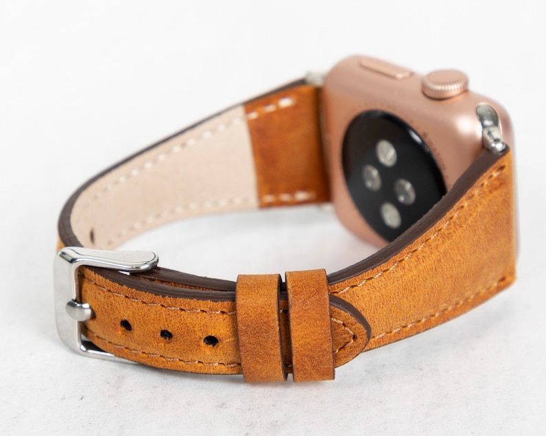 May include: A brown leather watch strap with a rose gold Apple Watch attached. The strap has white stitching detail and a silver buckle.