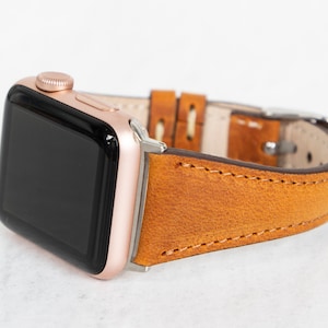 May include: A rose gold Apple Watch with a brown leather strap. The strap has a stitched edge and a buckle fastening.