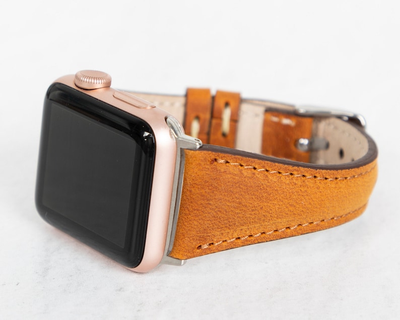 May include: A rose gold Apple Watch with a brown leather strap. The strap has a stitched edge and a buckle fastening.