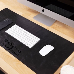 May include: A black leather desk mat with the text "Don't give up your dreams!"  The mat is on a wooden desk with a computer monitor, keyboard, mouse, phone, and a vase of pink flowers.