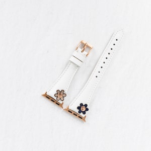 Pode incluir: Duas pulseiras de rel&oacute;gio de couro branco com fechos dourados e detalhes de flores. Uma pulseira tem uma flor branca com um centro dourado, e a outra tem uma flor azul com um centro dourado.