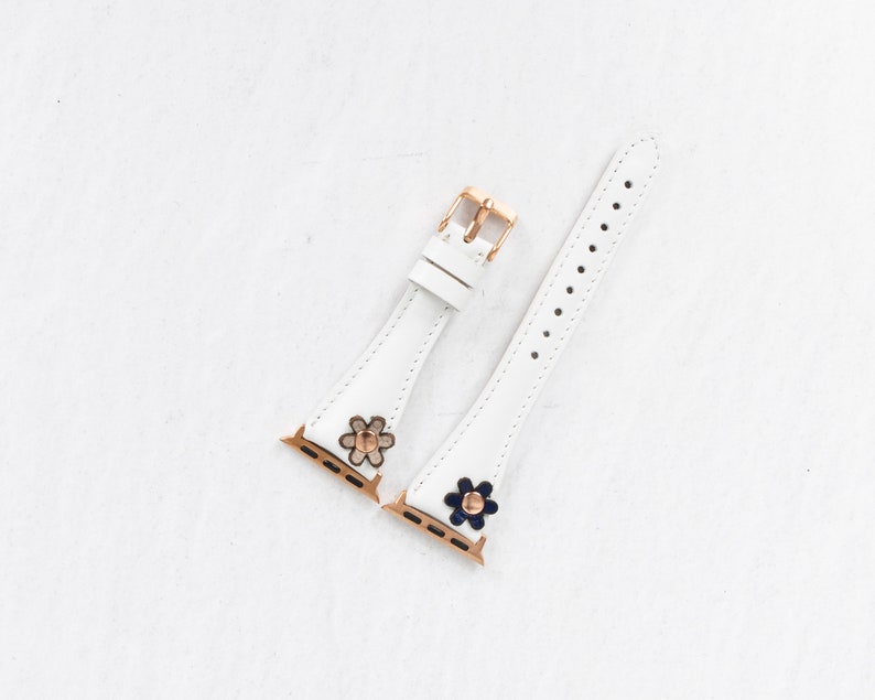 Pode incluir: Duas pulseiras de rel&oacute;gio de couro branco com fechos dourados e detalhes de flores. Uma pulseira tem uma flor branca com um centro dourado, e a outra tem uma flor azul com um centro dourado.