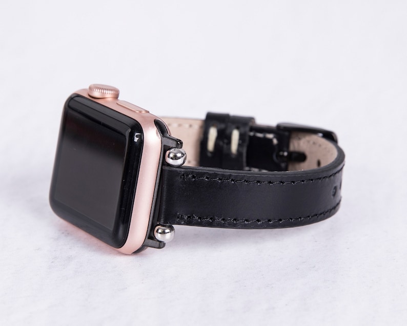 Puede incluir: Un Apple Watch de oro rosa con una correa de cuero negro. La esfera del reloj es negra y la correa tiene un borde cosido.