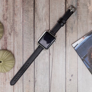 Puede incluir: Una correa de reloj de cuero negro con una esfera de reloj plateada. El reloj est&aacute; sobre una superficie de madera.