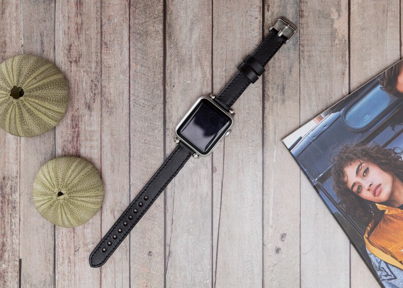 Puede incluir: Una correa de reloj de cuero negro con una esfera de reloj plateada. El reloj est&aacute; sobre una superficie de madera.