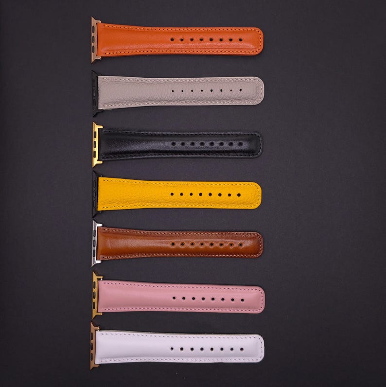 K&ouml;nnte beinhalten: Sieben Leder-Uhrenarmb&auml;nder in verschiedenen Farben, darunter Orange, Grau, Schwarz, Gelb, Braun, Rosa und Wei&szlig;. Jedes Band hat eine Reihe von L&ouml;chern zur Anpassung und wird vor einem dunklen Hintergrund gezeigt.