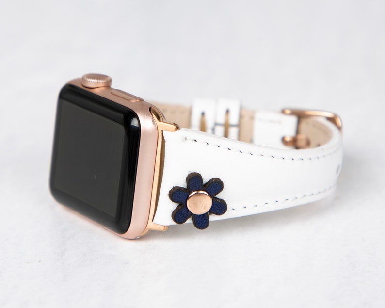 Pode incluir: Pulseira de rel&oacute;gio Apple de couro branco com fivela de ouro rosa e um detalhe de flor azul marinho.